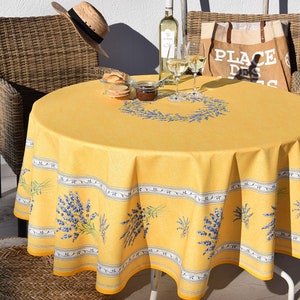 May include: A round yellow tablecloth with a lavender design, set on a patio table. The tablecloth features a border with lavender sprigs and a central lavender wreath. A straw hat and wine glasses are also visible.