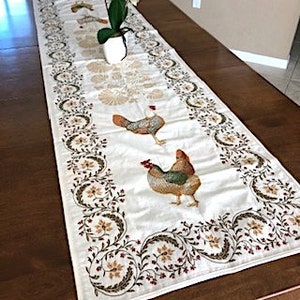 May include: A white table runner with a floral border and embroidered chickens. The chickens are brown, green, and red.