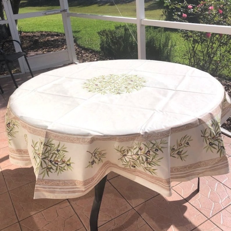 Puede incluir: Un mantel redondo blanco con un dise&ntilde;o de ramas de olivo verdes. El mantel tiene un borde beige con un patr&oacute;n repetido de ramas de olivo. El mantel est&aacute; puesto sobre una mesa redonda con una base de metal.