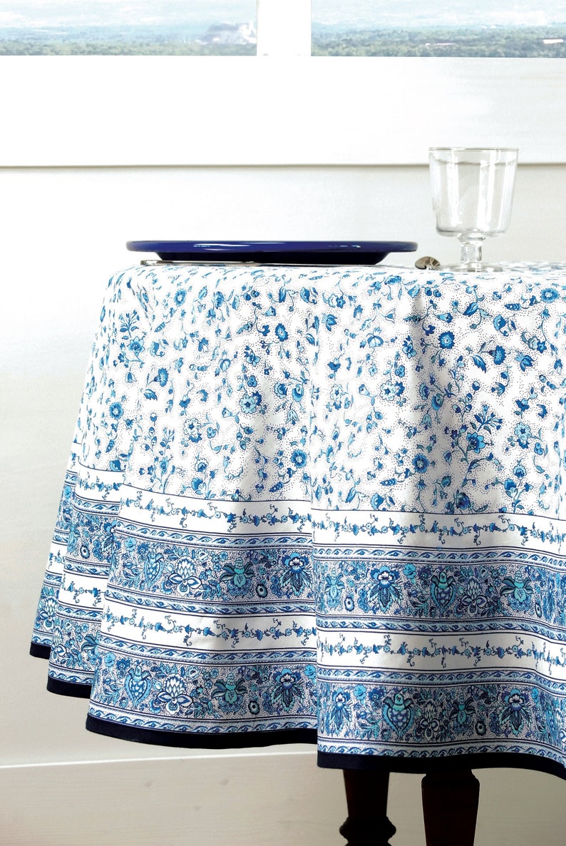 May include: A round tablecloth with a white background and a blue floral pattern. The tablecloth has a decorative border with blue floral and paisley designs. A blue plate and a clear glass sit on the table.
