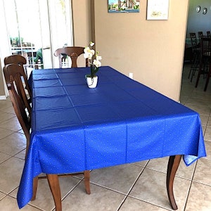 May include: A blue tablecloth with white polka dots covers a wooden dining table with four chairs. A small white vase with white flowers sits in the center of the table.