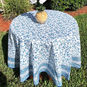 May include: A round tablecloth with a white background and a blue floral pattern. The tablecloth has a blue border and is draped over a wooden table. A glass vase with yellow filler and white flowers sits on top.