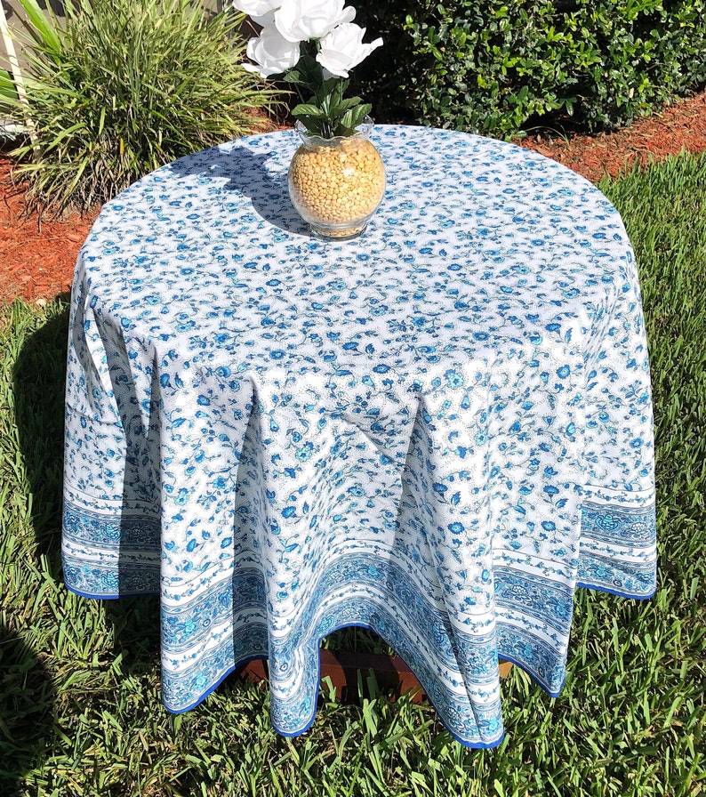 May include: A round tablecloth with a white background and a blue floral pattern. The tablecloth has a blue border and is draped over a wooden table. A glass vase with yellow filler and white flowers sits on top.