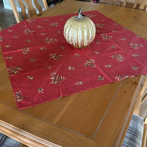 May include: A decorative glass pumpkin with a silver stem sits on a red tablecloth with olive branch patterns. The tablecloth is on a wooden table, surrounded by wooden chairs. The pumpkin is gold and has a textured surface.