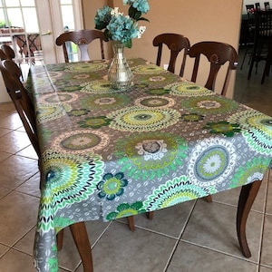 Pode incluir: Uma toalha de mesa estampada em cinza e verde com um design floral. A toalha de mesa está posta sobre uma mesa de madeira com cadeiras ao redor. Um vaso com flores azuis fica no centro da mesa.