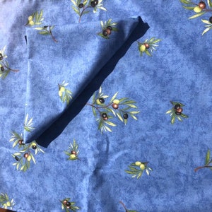 May include: A blue tablecloth with a pattern of olive branches and olives. The tablecloth is folded in half and has a napkin on top of it.