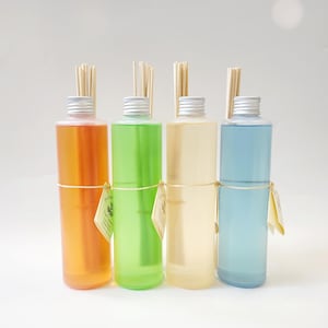 May include: Four glass bottles with reed diffusers, each filled with a different colored liquid: orange, green, yellow, and blue. The bottles have silver caps and are arranged in a row on a white surface.