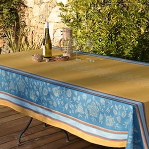 May include: A rectangular mustard-colored tablecloth with a blue floral border. A bottle of wine, two wine glasses, and a small bowl are on the table. The table is set outdoors on a wooden deck.