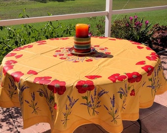 May include: A round yellow tablecloth with a floral design featuring red poppies and lavender sprigs. A candle sits on a decorative plate in the center of the table. The tablecloth is on a black metal table.