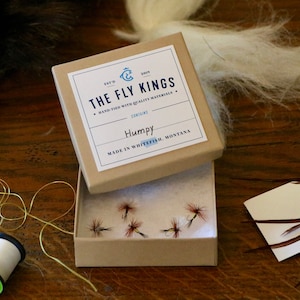 May include: A small, open cardboard box containing several artificial fishing flies labeled "Humpy." The box has a label with the text "THE FLY KINGS" and "MADE IN WHITEFISH, MONTANA." Spools of gold and white thread and feathers are visible.
