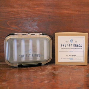 May include: A clear plastic fly box and a tan box with the text "THE FLY KINGS" and "Six Dry Flies." The fly box has a black hinge and contains vertical dividers. The tan box has a blue logo and text.