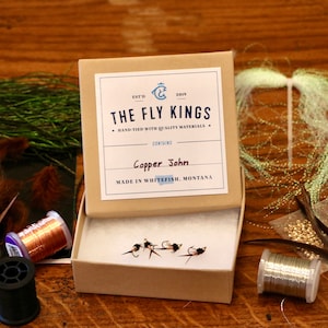 May include: A small, open cardboard box containing several Copper John fishing flies. The box has a label that reads "THE FLY KINGS" and "HAND-TIED WITH QUALITY MATERIALS." The flies are black and copper-coloured. Other fly-tying materials are visible.