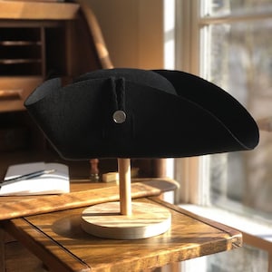 May include: A black felt tricorn hat with a silver button on the side, displayed on a wooden stand.