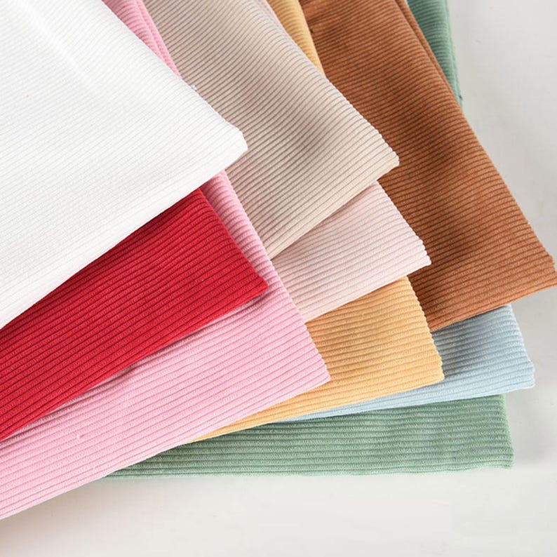 May include: A collection of folded fabric swatches featuring a ribbed texture. The colors included are white, red, light pink, beige, tan, brown, yellow, light blue, and sage green. Each piece of fabric is layered to show the vertical ribbing.