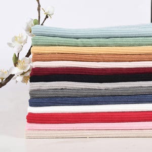 May include: A stack of folded ribbed fabric pieces in various colors including light blue, sage green, yellow, tan, cream, burgundy, black, gray, dark blue, white, red, pink, and beige, positioned next to a branch with white flowers.
