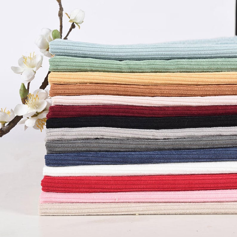 May include: A stack of folded ribbed fabric pieces in various colors including light blue, sage green, yellow, tan, cream, burgundy, black, gray, dark blue, white, red, pink, and beige, positioned next to a branch with white flowers.