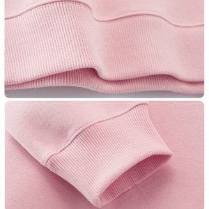 May include: Close-up of a light pink sweatshirt. The image highlights the ribbed cuffs and hem of the garment, showcasing the texture and stitching details. The fabric appears soft and comfortable, suggesting a casual style.