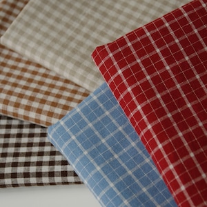 Puede incluir: Primer plano de varias muestras de tela en varios colores. Las telas presentan un patrón de cuadros clásico en tonos de rojo, azul, marrón, beige y marrón oscuro. Los cuadrados están formados por líneas blancas.