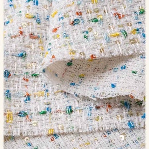 May include: White fabric with a woven texture and colorful flecks throughout. The fabric is folded over itself, showing the underside.