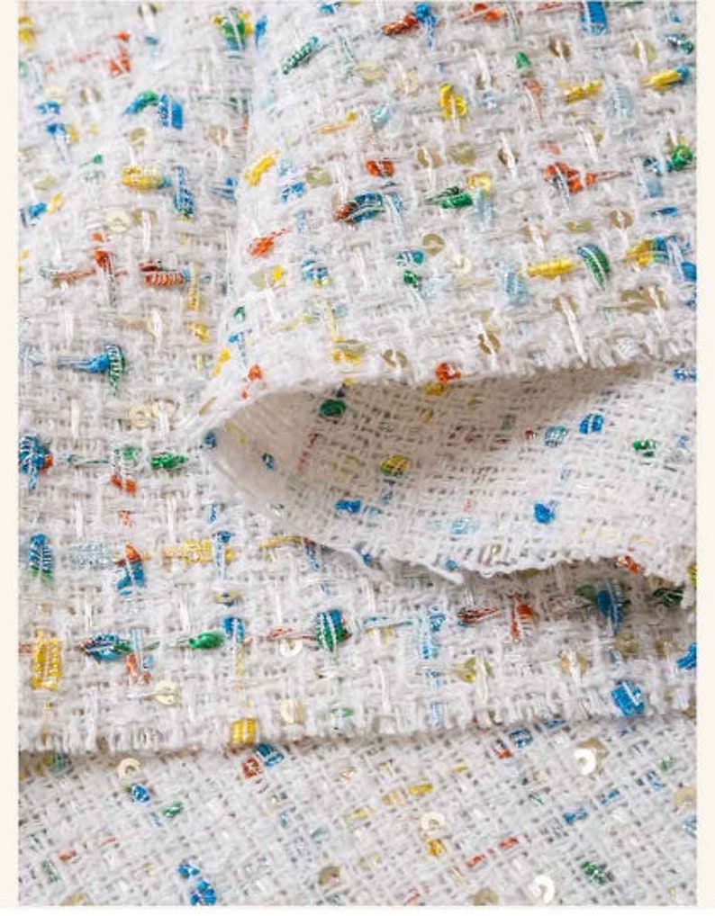 May include: White fabric with a woven texture and colorful flecks throughout. The fabric is folded over itself, showing the underside.