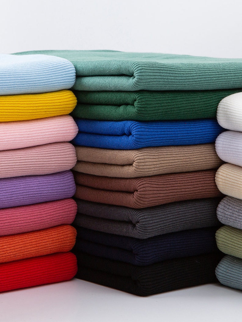 May include: A stack of folded ribbed knit fabric in various colors. The colors include light blue, yellow, pink, purple, orange, red, green, blue, brown, black, and gray. The fabric appears soft and has a textured surface.