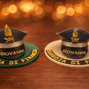 Può includere: Due cappelli blu navy in miniatura con emblemi dorati, ciascuno su una base circolare. Una base è verde, l'altra bianca, entrambe con le parole "GIOVANNI" e "GUARDIA DI FINANZA" in oro. Lo sfondo è sfocato con bokeh caldo.