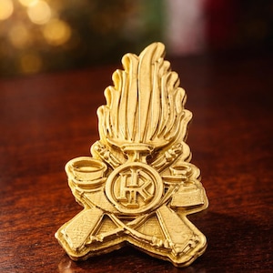 Puede incluir: Un emblema dorado con una llama estilizada en la parte superior, herramientas cruzadas y un diseño circular central. El emblema es probablemente una insignia o un pin, con detalles intrincados y un acabado pulido.