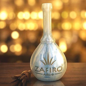 Può includere: Una bottiglia di vetro trasparente a forma di pera con la scritta "ZAFIRO ANEJO" in lettere dorate. La bottiglia ha un collo lungo e un tappo di legno con un nappa. Lo sfondo è sfocato con toni caldi e dorati.