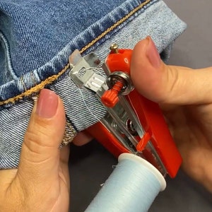 May include: A red and silver handheld sewing machine with a spool of light blue thread. The machine is being used to sew the hem of a pair of blue jeans.