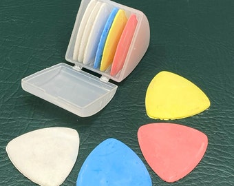 May include: A set of colorful tailor's chalk in a clear plastic case. The chalk pieces are white, blue, yellow, and red, and are triangular in shape. Three loose pieces of chalk are also visible. The background is a dark green surface.