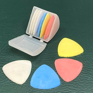 May include: A set of colorful tailor's chalk in a clear plastic case. The chalk pieces are white, blue, yellow, and red, and are triangular in shape. Three loose pieces of chalk are also visible. The background is a dark green surface.