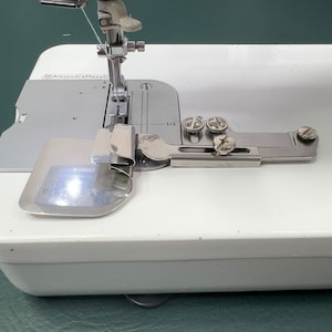 May include: Close-up of a white sewing machine with a silver foot pedal and a silver needle. The machine is set up for sewing with a white thread.