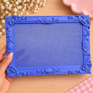 May include: A rectangular blue display board featuring an ornate, decorative frame with intricate scrollwork and shell-like motifs. The center of the board consists of a fine, uniform grid pattern, providing a structured surface for organizing items.