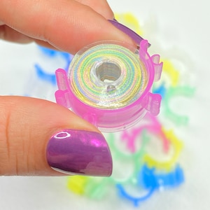 May include: A clear plastic sewing machine bobbin filled with iridescent thread. The bobbin is held between fingers, with a purple nail polish visible. Other colorful plastic items are in the background.