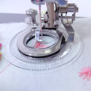 May include: Close-up of a sewing machine needle and foot, with a clear plastic hoop and a pink thread. The machine is stitching a pink flower design on white fabric.