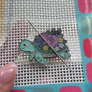 May include: A decorative turtle-shaped needlepoint canvas embellishment with a floral design. The turtle is light green with a pink belly and is adorned with a variety of colorful flowers. A silver needle is inserted through the canvas.