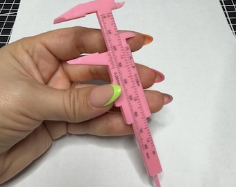 Könnte beinhalten: Ein rosa Kunststoff-Messschieber mit Millimeter- und Zollmessungen. Das Werkzeug wird vor einer weißen Oberfläche gehalten. Der Messschieber hat eine verschiebbare Skala und eine Spitze, für präzise Messungen.