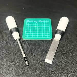 May include: Two white handled tools with black accents, one with a pointed tip and the other with a flat blade, on a black surface with a green cutting mat labeled "Cutting Mat".