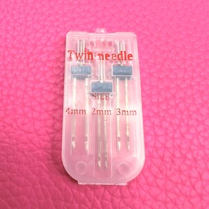 May include: A clear plastic case containing three twin needles for sewing. The case has the text "Twin needle" and "SIZE" printed on it. The needles are labelled with sizes 1mm, 2mm, and 3mm.