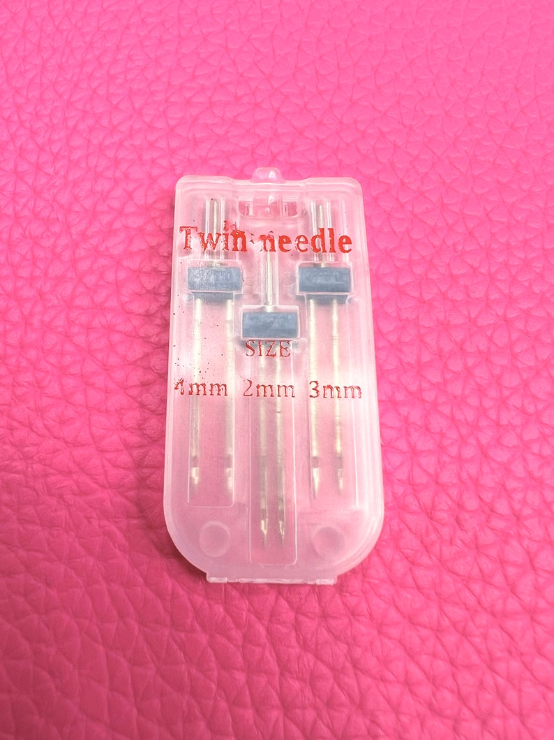 May include: A clear plastic case containing three twin needles for sewing. The case has the text "Twin needle" and "SIZE" printed on it. The needles are labelled with sizes 1mm, 2mm, and 3mm.