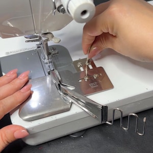 May include: A close-up of a sewing machine with a metal attachment. The machine is white with silver metal components. A hand is holding a metal tool near the attachment. The text "DYLT 30mm" is visible on the attachment.