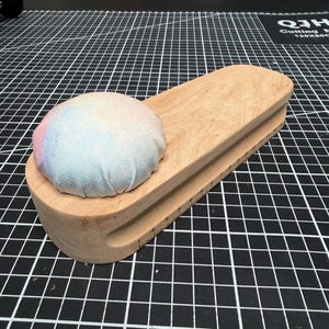 May include: A wooden tailor's clapper with a rounded, multi-colored pin cushion attached. The clapper is light-colored wood with a ruler etched along one side. The pin cushion has pastel colors of pink, blue, and yellow.