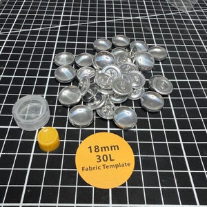 May include: A collection of silver-colored, domed buttons scattered on a black and white grid. Also present are a clear plastic container, a yellow button, and a round orange label with the text "18mm 30L Fabric Template".