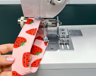 Könnte beinhalten: Eine weiße Nähmaschine mit einem rosa Stoff mit Erdbeerprint, der genäht wird. Der Stoff wird von einer Hand gehalten.