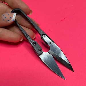 May include: Small, silver-bladed thread snips with black handles. The word "EAGLE" is printed on the handle. The snips are open, ready for use, and are set against a bright pink background.