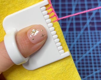 May include: A close-up of a craft project in progress. Bright yellow felt is being sewn with pink thread. A white plastic tool with a finger loop is used to guide the needle. The background is a blue grid.