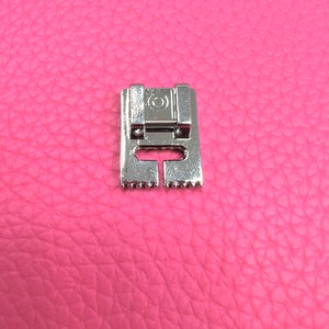 May include: A silver sewing machine foot with a small circle on the top and a row of teeth on the bottom.
