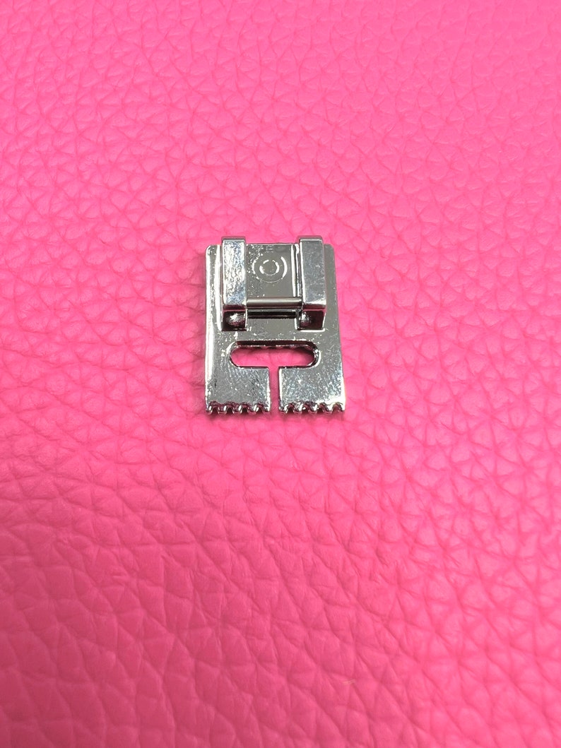 May include: A silver sewing machine foot with a small circle on the top and a row of teeth on the bottom.