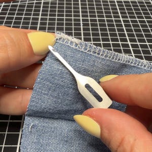 May include: A silver-colored metal bodkin tool with a flat, rectangular handle and a pointed tip with a small eyelet, positioned against a piece of blue denim fabric that features a white serged edge finish along the top seam.