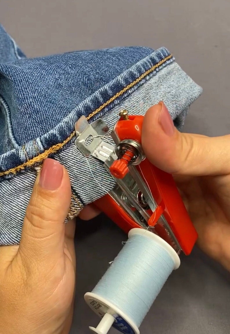 K&ouml;nnte beinhalten: Eine rote und silberne Handn&auml;hmaschine mit einer Spule hellblauen Fadens. Die Maschine wird verwendet, um den Saum einer blauen Jeans zu n&auml;hen.